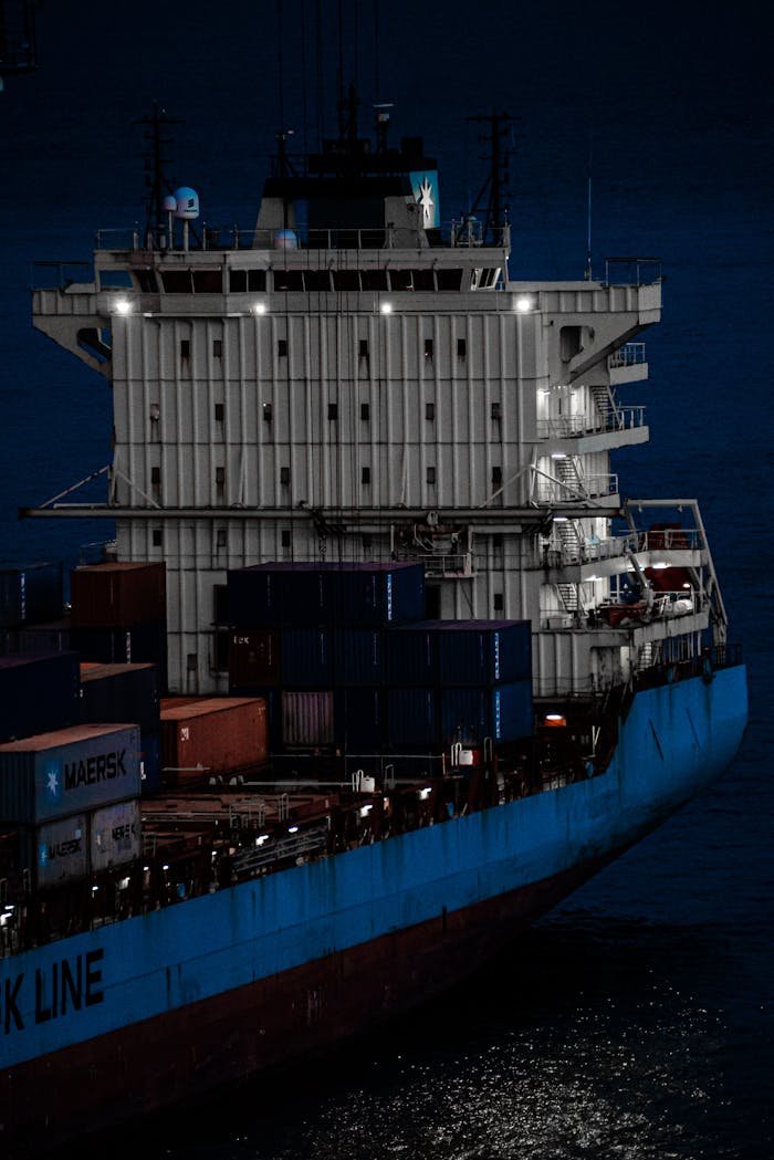 A large container ship sailing on the ocean at night, illuminated by lights.